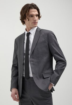 Giacca da suit grigia con vestibilità su misura, chiusura a un bottone, revers a scollo e taschino sul lato sinistro del petto; abbinata a una camicia bianca e cravatta nera.