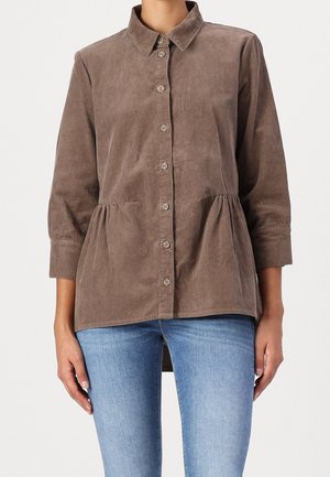 Brązowa koszula sztruksowa zapinana na guziki z marszczonym detalem w talii, rękawy do trzy czwarte, noszona z jasnoniebieskimi dżinsami.