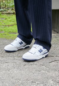 Zapatilla blanca y gris con acentos en azul marino, materiales de cuero texturizado y malla, con un prominente logo de la N y una suela de goma.