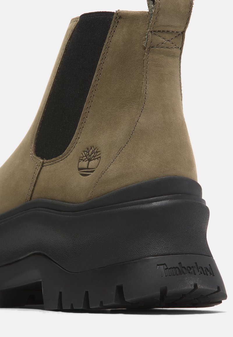 Olijfgroene suède enklaarzen met zwarte elastische zijpanelen en een gestructureerde zwarte rubberen zool. Kenmerkt het reliëf Timberland-logo.