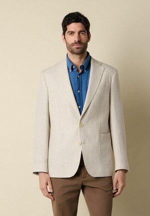 Man met donker haar en baard, gekleed in een beige geruite blazer, een blauw overhemd met knopen en een bruine broek, staand tegen een beige achtergrond.