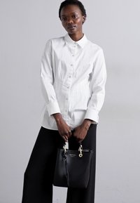 Camicia bianca con colletto, abbinata a pantaloni neri a gamba larga. Una borsa nera presenta dettagli in oro. Tessuto dalla texture liscia.