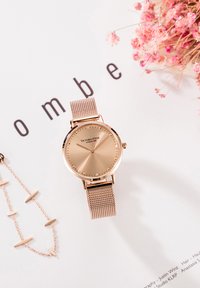 Orologio da polso in rete oro rosa con quadrante semplice accanto a una catena delicata coordinata e fiori secchi rosa su superficie bianca con testo nero.