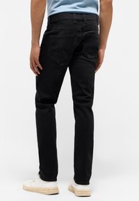 Czarne dżinsy o prostym kroju. Cechy to tylne kieszenie i szlufki na pasek. Noszone z białymi sneakerami, widok z tyłu.