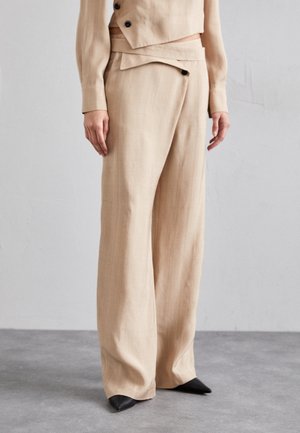 Beige maßgeschneiderte Hose aus leichtem Material, mit weitem Bein, flachem Frontbund und schwarzen spitzen Schuhen.