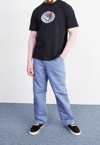 Zwart katoenen T-shirt met afbeelding van een flesdop; lichtblauwe broek met een losse pasvorm; zwarte sneakers met witte veters; binnenomgeving.