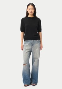 Kvinna med långt mörkt hår som bär en svart kortärmad tröja och ljusblå slitna jeans med vida ben samt sandaler, stående mot en vit bakgrund.