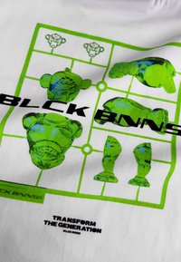 Weißes T-Shirt mit einem grünen grafischen Design stilisierter Spielzeugfiguren, mit fettem Text "BLCK BNNS" und "TRANSFORM THE GENERATION" darunter.