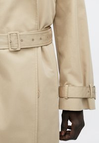 Trench-coat beige avec ceinture assortie attachée à la taille et sangle au niveau du poignet, porté sur le bras et la main d'une personne.