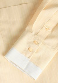 Manchette de chemise à manches longues rayée jaune et blanche avec deux boutons jaunes et une poche sur un fond en tissu rayé jaune.