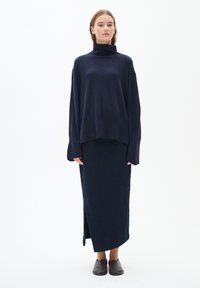 Modra turtleneck pulover s dolgimi rokavi, v kombinaciji z ujemajočo se dolgo, teksturirano krilo z bočnim razporkom, dopolnjeno z temnimi natikači.