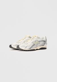 Baltas un pelēks New Balance sporta apavi ar krēmkrāsas šņorēm, melniem zoliem un sieta paneļiem, novietoti blakus uz balta fona.