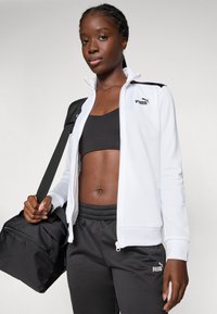 Vit zip-up jacka med en logotyp, bärs över en svart crop top. Svarta träningsbyxor med elastisk midja. Håller en svart duffelväska.