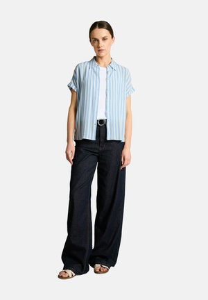 Frau steht und trägt ein kurzärmeliges blau-weiß gestreiftes Hemd mit Knöpfen, ein weißes Top, schwarze Hose mit weitem Bein, schwarzen Gürtel und beigen Sandalen.