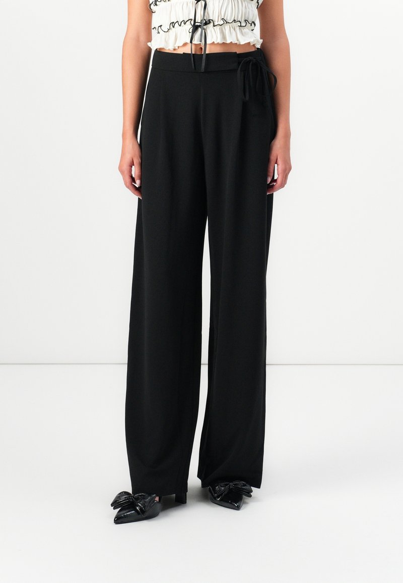 Femme portant un pantalon noir taille haute à jambes larges avec un détail de lien sur le côté, associé à des chaussures noires pointues avec des accents de nœuds.