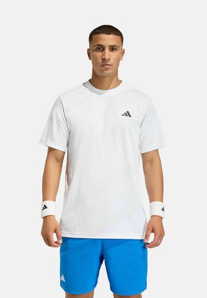 Jonge man die een wit Adidas T-shirt, witte polsbandjes en blauwe Adidas short draagt, staand tegen een effen lichte achtergrond.