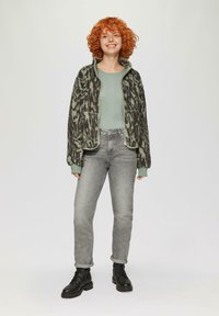 Polarowa kurtka w zielono-czarny wzór, prążkowany jasnozielony sweter, szare dżinsy oraz czarne buty na sznurówki, stojąca na jasnym tle.