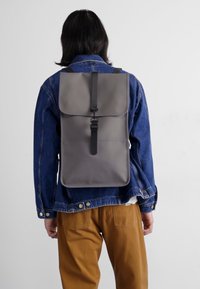 Zaino grigio con patta pieghevole, chiusura a fibbia e finitura opaca, indossato sopra una giacca in denim, dotato di spallacci regolabili.