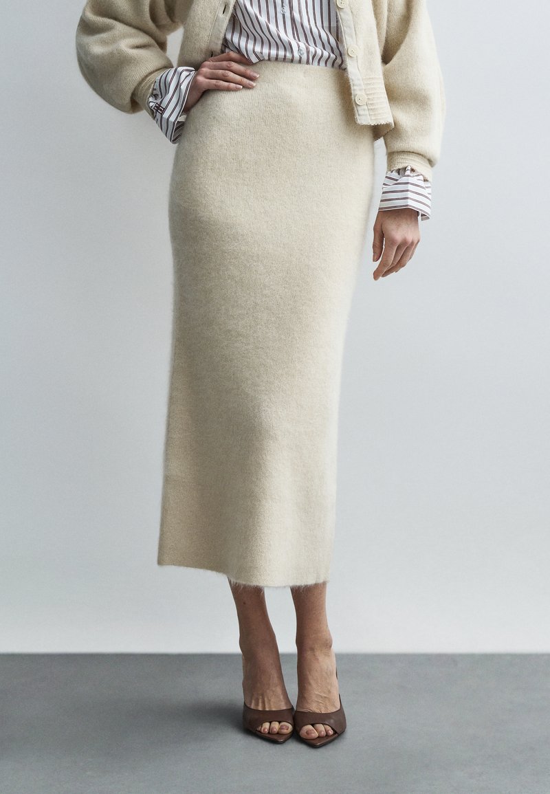 Moteris dėvi kremiškos vilnos sijoną, dryžuotą marškinių palaidinę, smėlio spalvos megztinį ir rudus atvirų pirštų aukštakulnius, stovinti priešais paprastą foną.