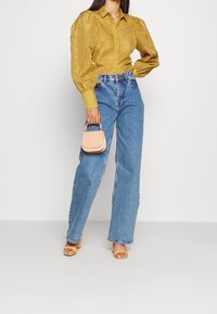 Blouse jaune moutarde à manches bouffantes et motif ajouré, assortie à un jean taille haute bleu clair et un petit sac à main rose.