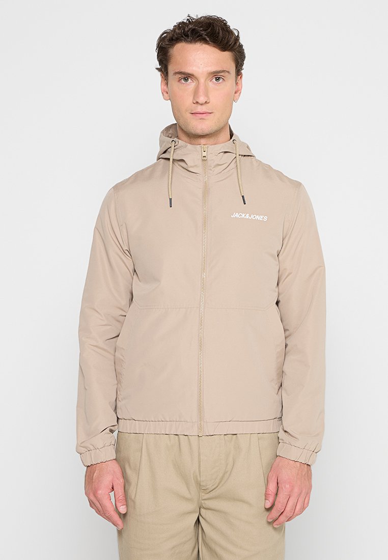 Chaqueta ligera beige con capucha, puños elásticos, cremallera frontal y detalles de logo. Textura suave con un diseño minimalista.