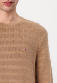 Beige neulepaita, jossa on vaakasuorat raidat, pyöreä kaula-aukko ja pieni sininen, valkoinen ja punainen brodeerattu logo rinnassa.
