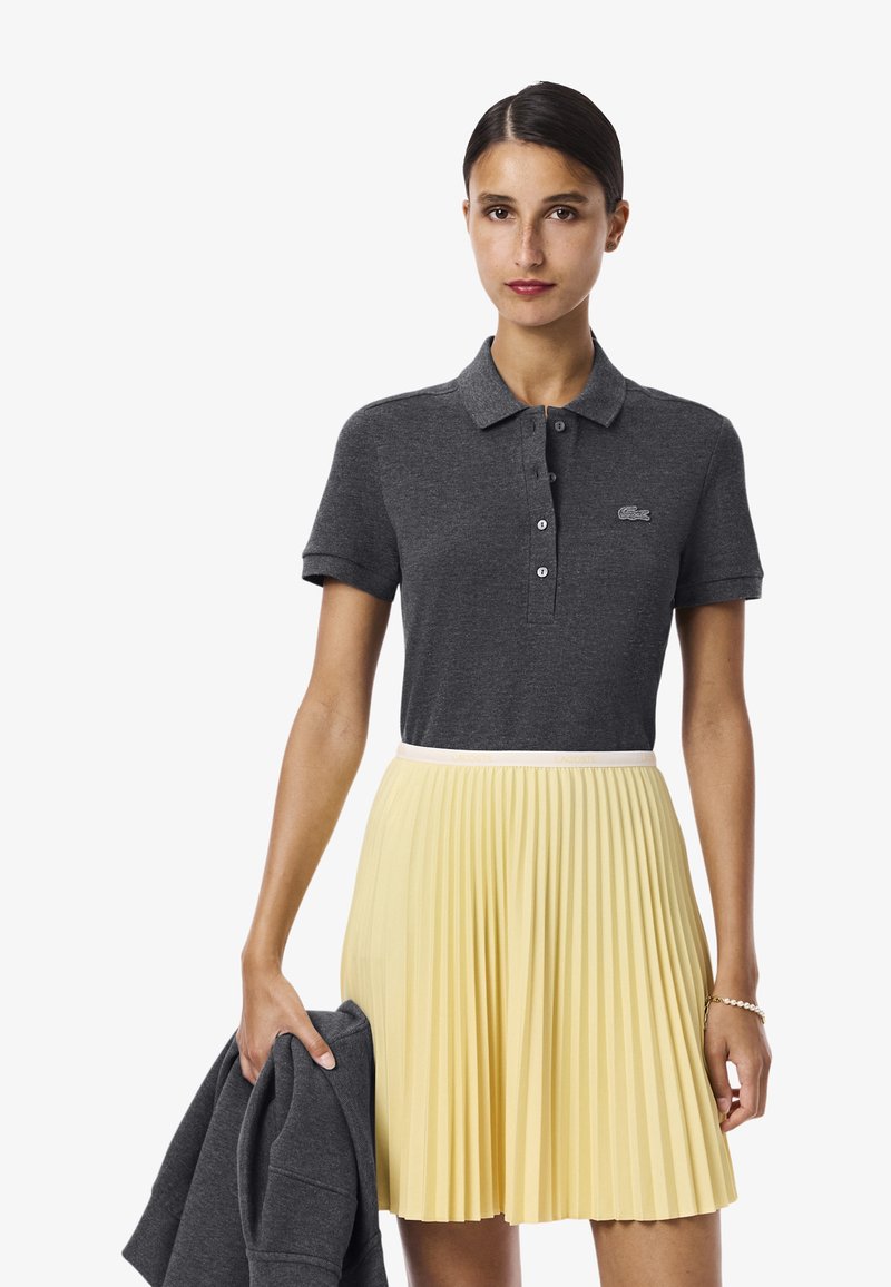 Grå polo-shirt med knappar, i kombination med en plisserad gul kjol. Modellen håller en mörkgrå sweatshirt. Släta texturer och kontrasterande färger.