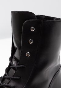 Botte en cuir noir avec une tige haute, surface texturée, trois œillets en argent et lacets noirs. Des détails de coutures mettent en valeur la structure.