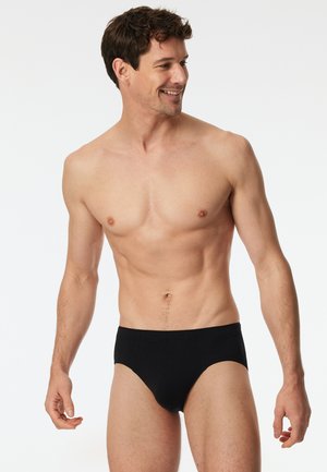 Schwarze Baumwollslips mit glatter Textur, elastischem Bund und figurbetontem Schnitt. Das Modell steht aufrecht vor einem neutralen Hintergrund.
