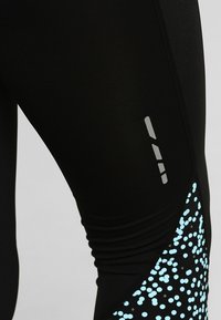 Primo piano di leggings atletici neri con pois azzurri chiari e strisce grigie riflettenti sulla coscia.