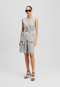 Grijze mouwloze jumpsuit met een ceintuur in de taille, zijzakken en een getailleerd ontwerp, gecombineerd met zwarte sandalen met bandjes en een zonnebril.