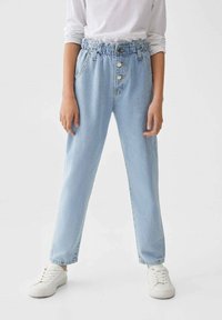 Jeans de mezclilla azul claro con cinturilla elástica, dos botones, bolsillos frontales y un corte relajado. Combinados con zapatillas blancas.