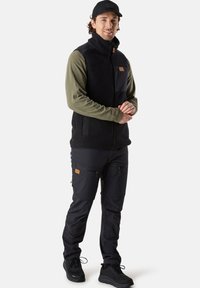 Svart fleeceväst över en olivgrön långärmad tröja, kombinerad med svarta cargo-byxor med dragkedjefickor och svarta sneakers.