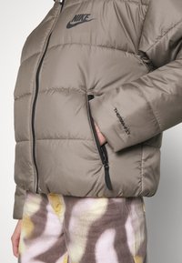 Graue Pufferjacke mit glänzender Oberfläche, ausgestattet mit einem schwarzen Logo auf der Brust und einer Reißverschlusstasche. Der Stoff hat eine gesteppten Textur.