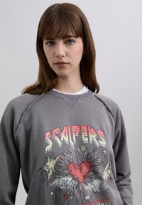 Gray crewneck sweatshirt with frayed neckline. Features a colorful graphic design with the word “SCAPERS” and heart motif.