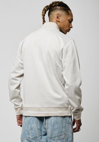 Chaqueta color beige claro con un patrón de cuadrícula texturizado, cuello y puños acanalados, que presenta un corte relajado y dobladillo recto. Combinada con pantalones cortos de mezclilla desgastados.