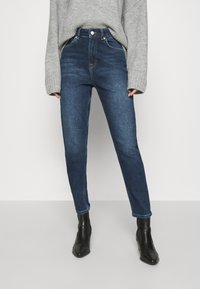 Donna che indossa jeans a vita alta blu scuro, un maglione grigio oversized e stivaletti neri, appoggiata a uno sfondo semplice.