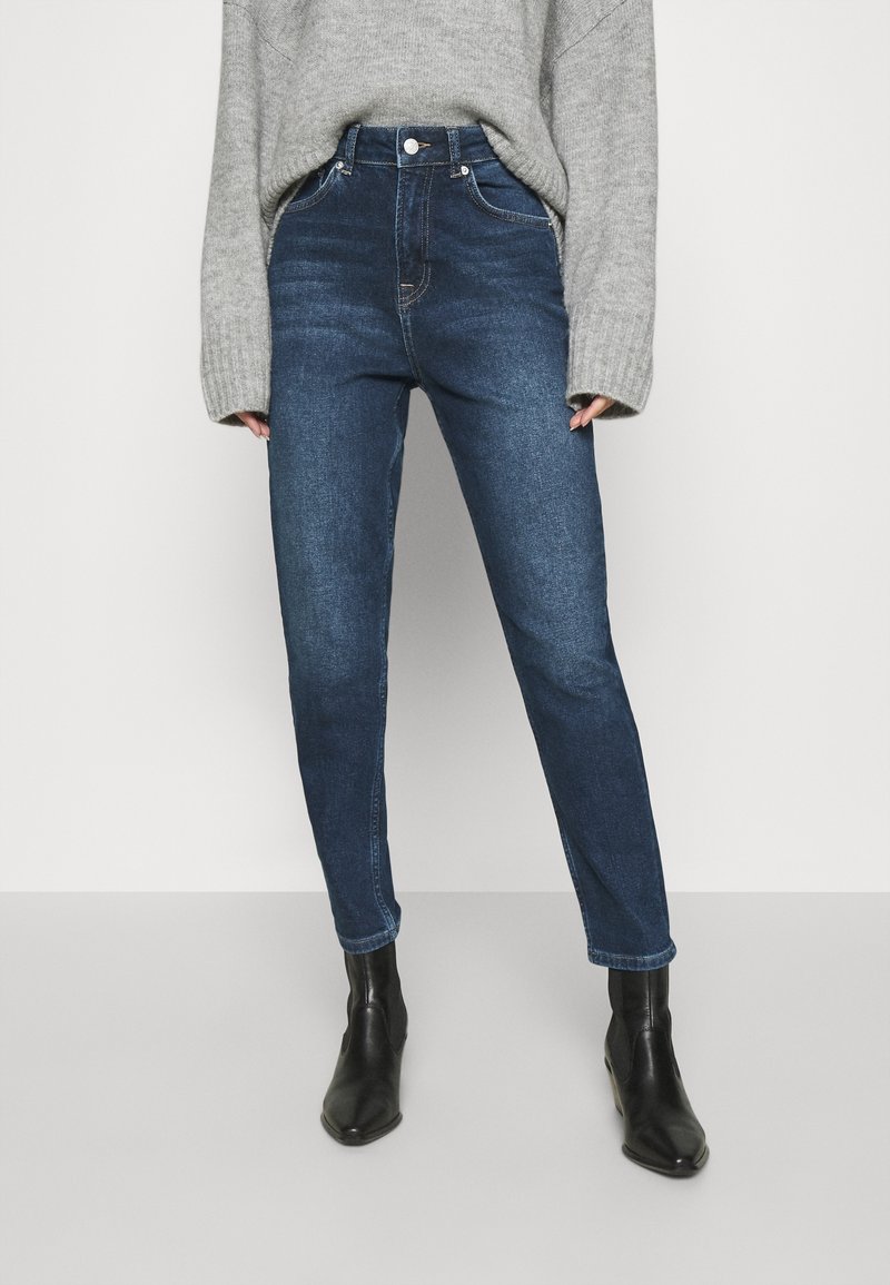 Donna che indossa jeans a vita alta blu scuro, un maglione grigio oversized e stivaletti neri, appoggiata a uno sfondo semplice.