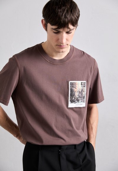Barna pamut póló, elülső zsebbel, amelyen nyomtatott városi táj látható. Rövid ujjak és kerek nyakú dizájn. Simább anyag.