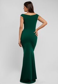 Vrouw in een lange, off-shoulder donkergroene jurk, staande met één hand op haar heup en van de camera afgekeerd.