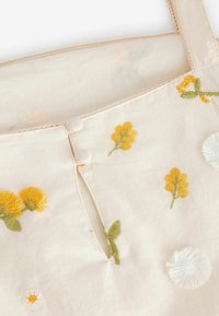 Top de tela bordada en beige claro con diseños florales amarillos y acentos blancos. Presenta botones y un delicado encaje en los tirantes.