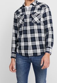 Chemise en flanelle à carreaux bleu marine et blanc, manches longues, deux poches poitrine et fermeture à boutons sur le devant. Texture douce et design avec ourlet arrondi.