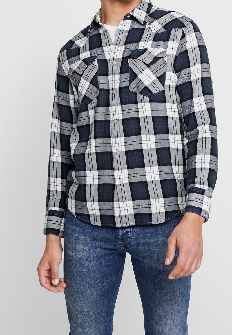 Chemise en flanelle à carreaux bleu marine et blanc, manches longues, deux poches poitrine et fermeture à boutons sur le devant. Texture douce et design avec ourlet arrondi.