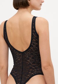 Body en dentelle noire avec un dos plongeant, présentant des motifs floraux doux et transparents ainsi que de fines bretelles. Finition lisse en noir le long des bords.