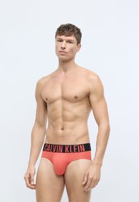 Červené bavlnené slipy s čiernym pásom s logom "CALVIN KLEIN". Dizajn je priliehavý a minimalistický, zdôrazňujúci tvar a formu.