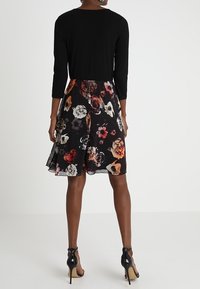 Schwarzes Oberteil mit langen Ärmeln, Rock mit floralem Muster in Rottönen, Rosa und Weiß, fließender Stoff, abgerundet mit schwarzen High Heels.