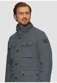Graue isolierte Jacke mit hohem Kragen, mehreren mit Knöpfen versehenen Klappen-Taschen und einem Logopatch auf der linken Schulter. Glatte Textur.