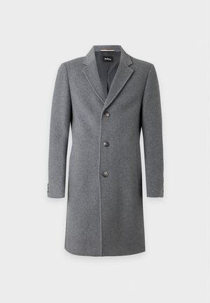 Homme debout portant un manteau gris boutonné sur un pantalon noir, un polo noir et des baskets noires sur un fond blanc uni.