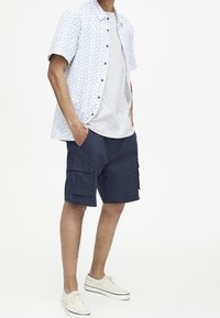 Kortærmet skjorte med knapper i hvid med blå mønstre, grå t-shirt og marineblå cargo shorts med flere lommer. Hvide sneakers.