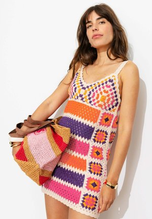 Femme portant une robe au crochet colorée avec des motifs géométriques, tenant un sac tissé multicolore, debout devant un fond blanc.